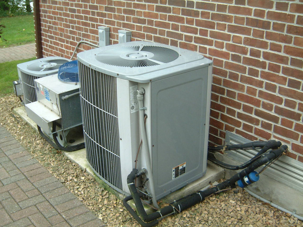 Outdoor HVAC units installed beside a brick wall, showcasing a cooling system with visible pipes and gravel landscaping, relevant to HVAC replacement discussions.