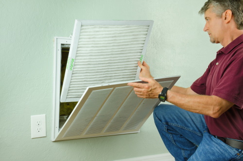 Man changing an AC filter in a wall vent, emphasizing the importance of regular maintenance for HVAC efficiency.