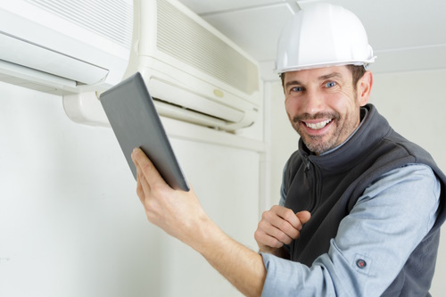 HVAC technician smiling while using a tablet near a wall-mounted cooling system, emphasizing the importance of updated HVAC regulations and sustainable refrigerants.